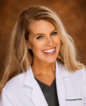 Clearwater Dental | Smiling woman with long blonde hair wearing a white coat and black turtleneck against a warm brown background